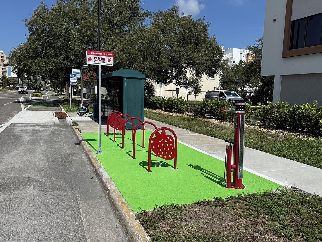 Green-surfaced bike hub with red strawberry-shaped racks and a cylindrical repair kiosk on a city sidewalk.