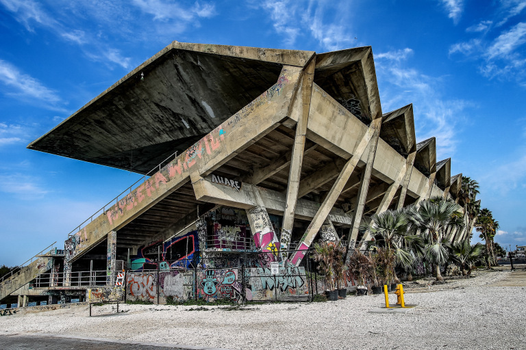 Abandoned concrete grandstand with a large cantilevered roof, covered in graffiti, surrounded by palm trees.