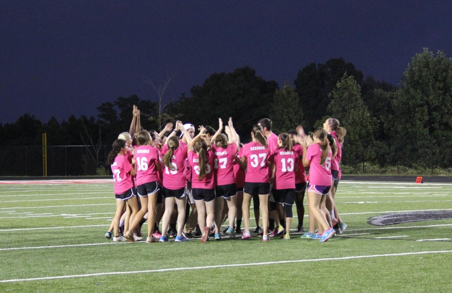 Young women in pink jerseys huddle together on a lit turf field at night.