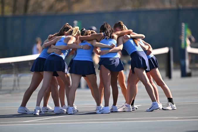 Female tennis players in light blue tops and navy skirts huddle together on an outdoor court.