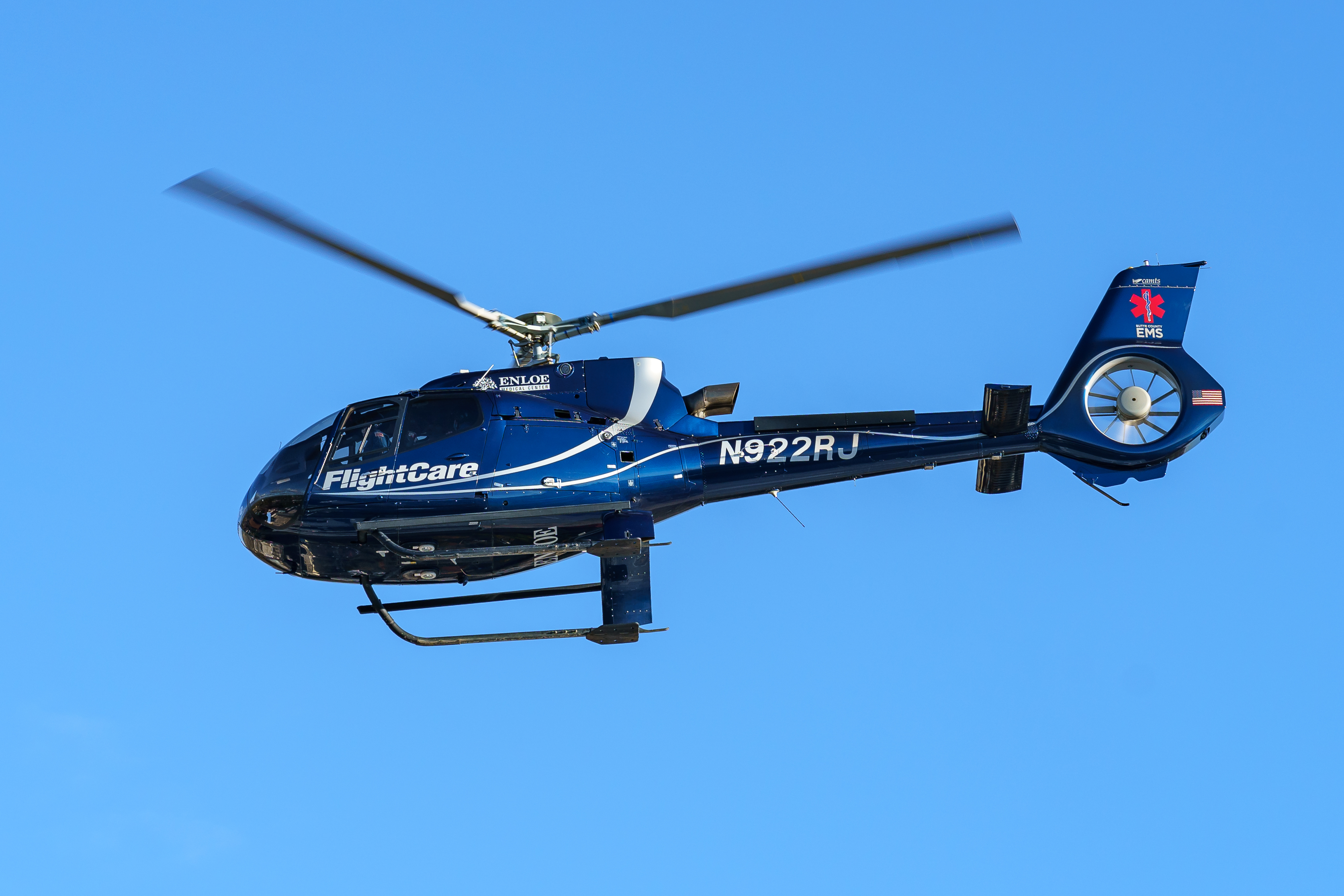 A dark blue medical helicopter with EMS star-of-life markings flies against a clear blue sky.