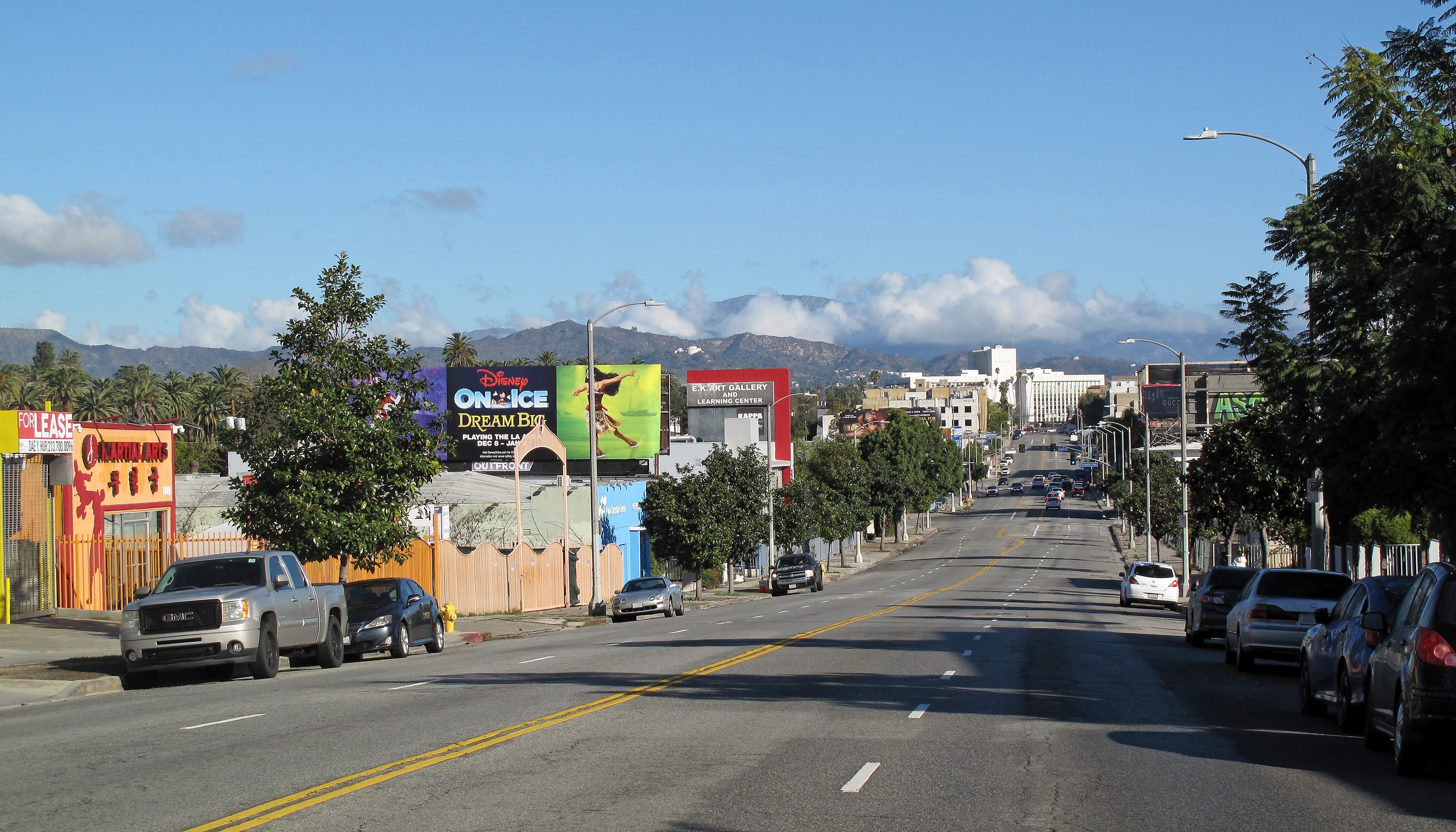 Wide urban boulevard with multiple lanes, tree-lined sidewalks, and mountains visible in the distance.