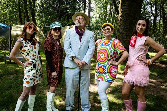 Five costumed actors pose outdoors in a wooded park, wearing colorful 1960s-inspired outfits.