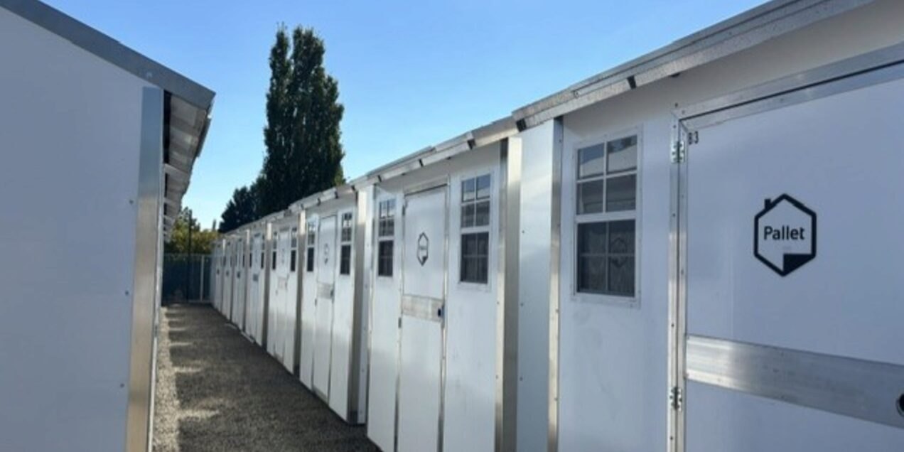 A row of white modular shelter pods with doors and windows along a gravel path.