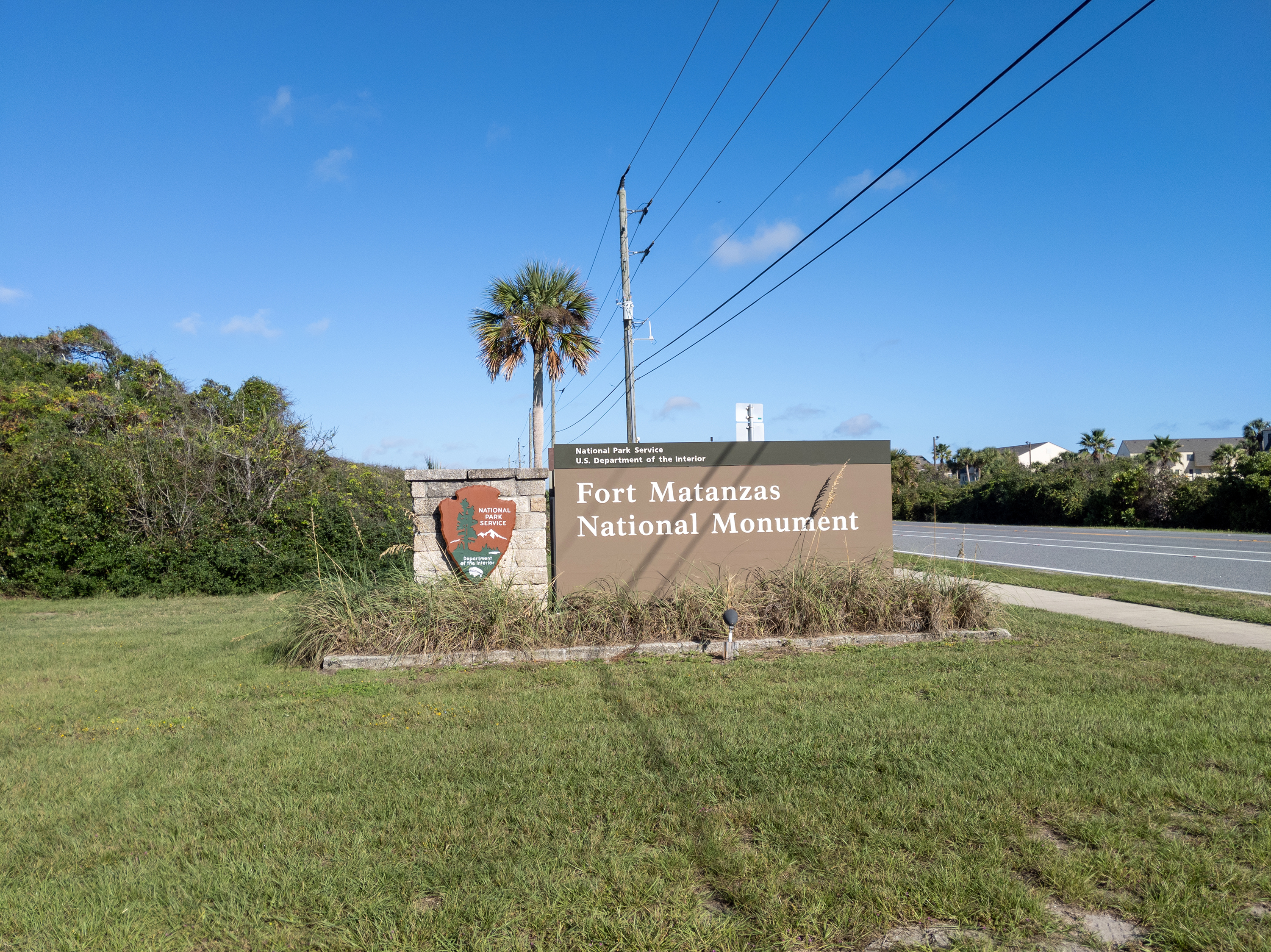 Roadside entrance sign for Fort Matanzas National Monument with palm trees and blue sky.