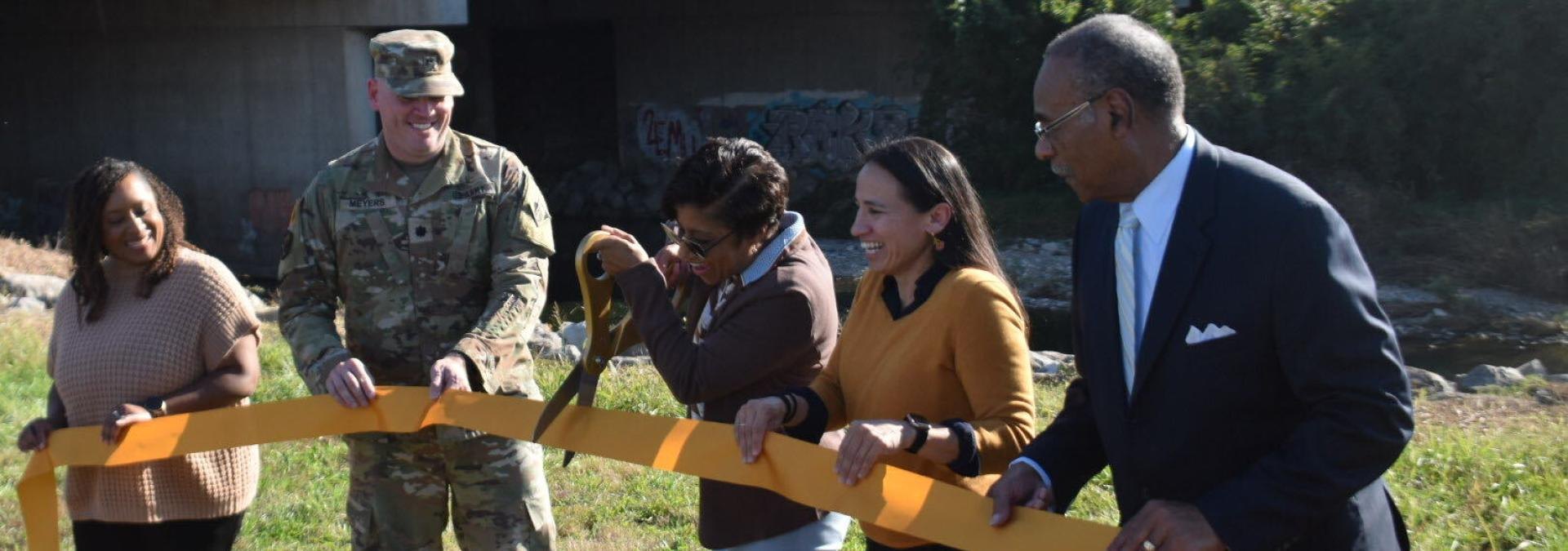 Five people cut an orange ribbon outdoors near a concrete flood control structure.
