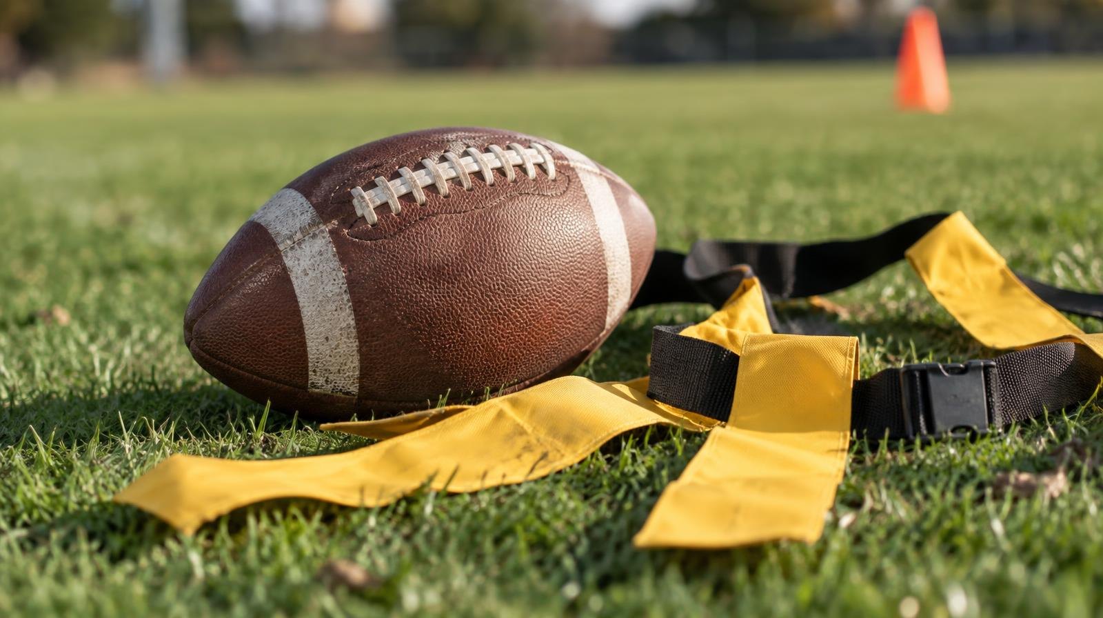A football with flag football belts and colorful flags laid on green grass.