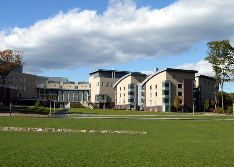 Wide green lawn stretching toward modern college buildings under a partly cloudy sky.