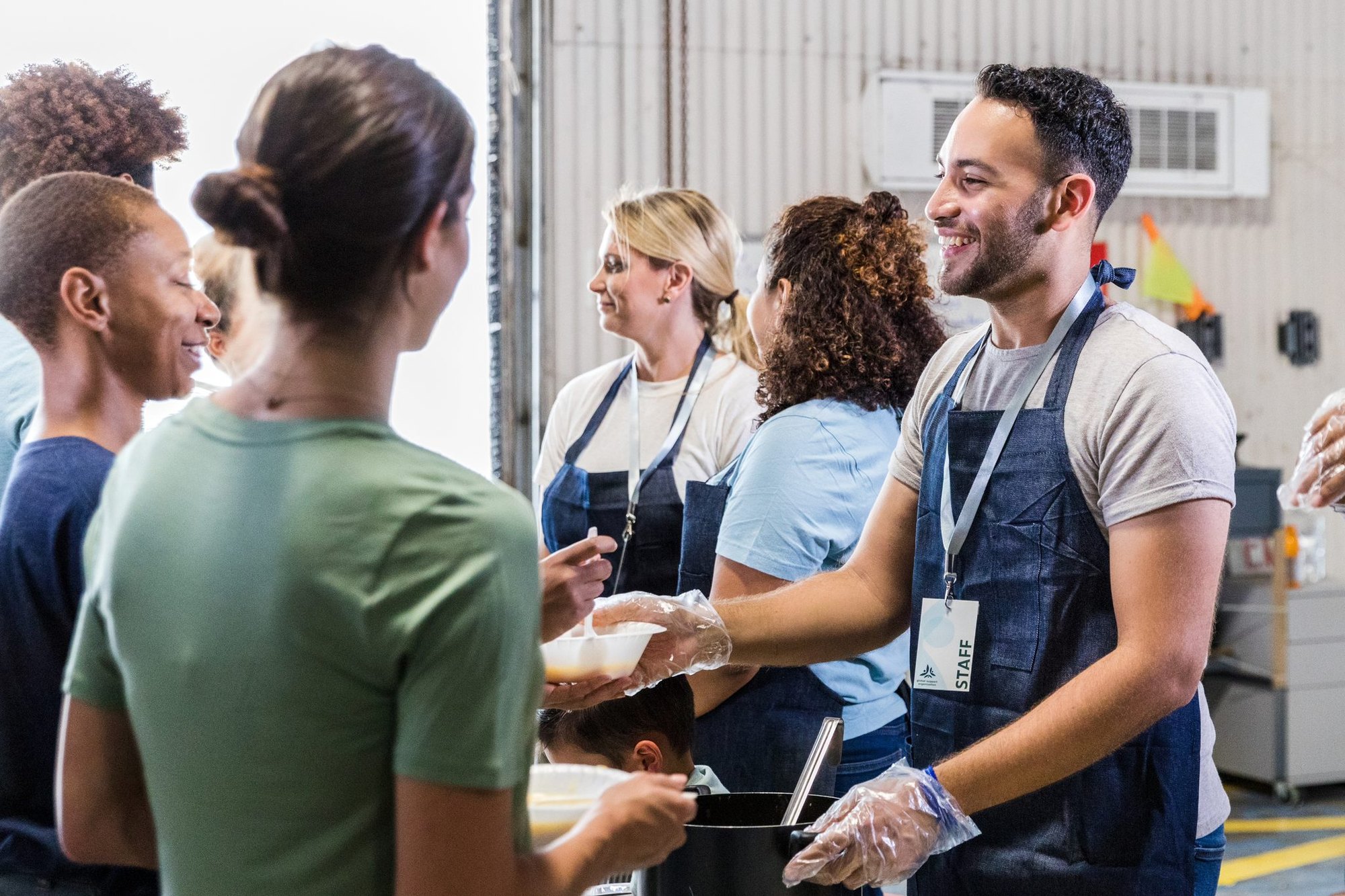 Volunteers in blue aprons serve food to people at an indoor food distribution site.
