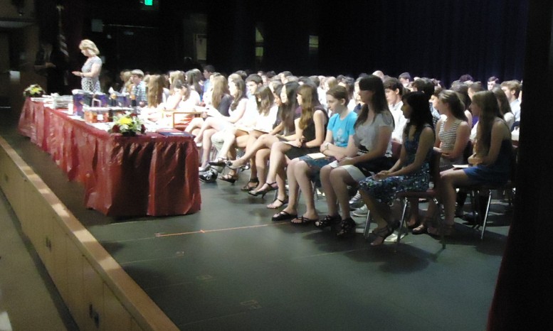 Students in formal attire seated in rows on a school auditorium stage.