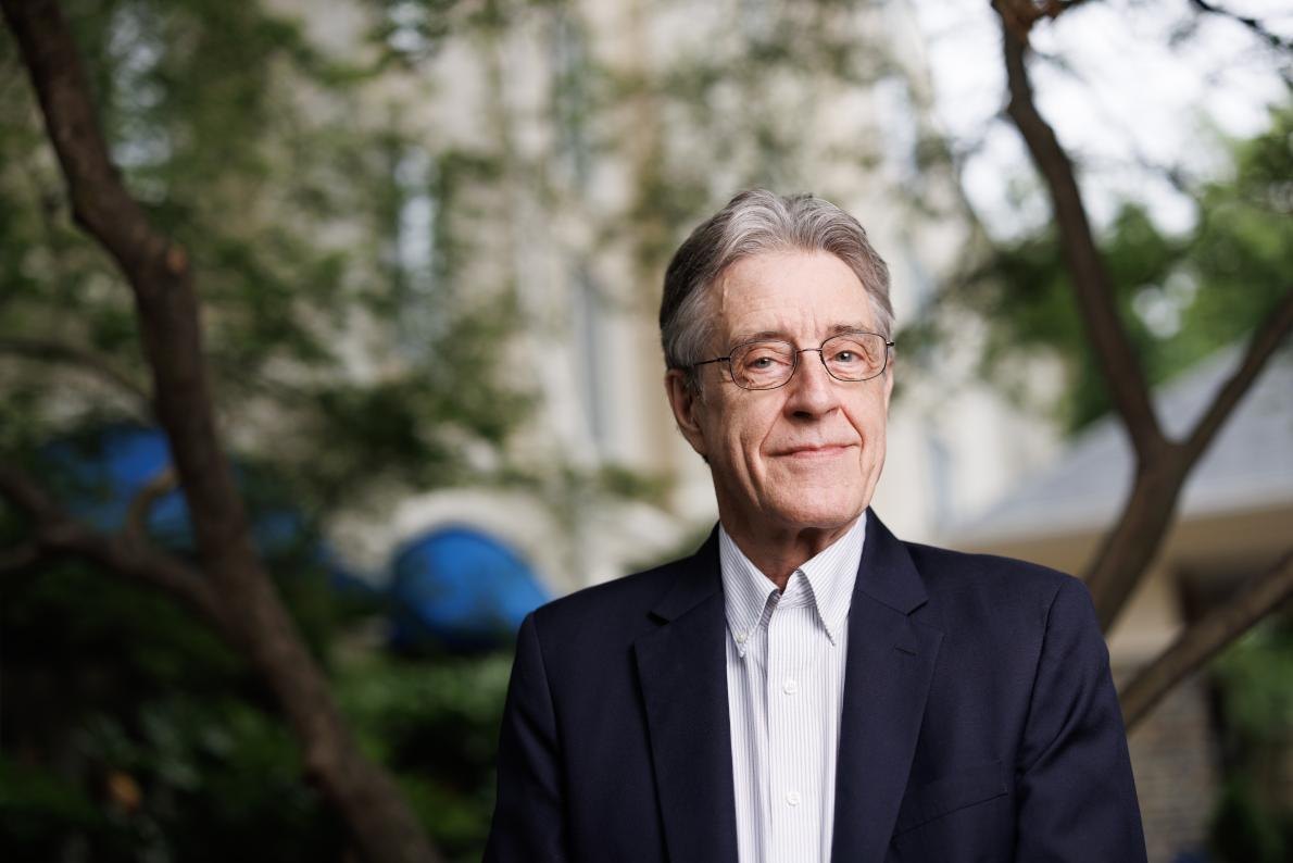 Older man with gray hair and glasses in a dark blazer and white shirt, smiling outdoors with trees behind him.