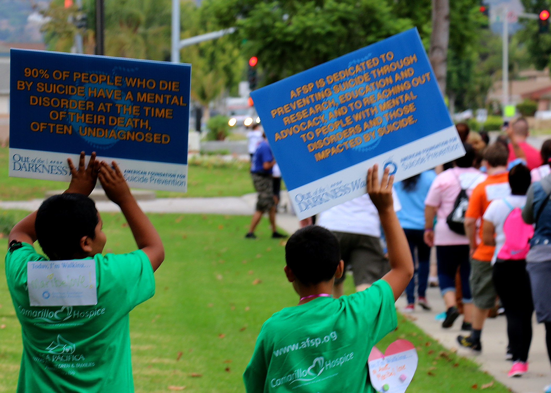 Two children in green shirts hold blue AFSP suicide prevention signs at an outdoor walk.
