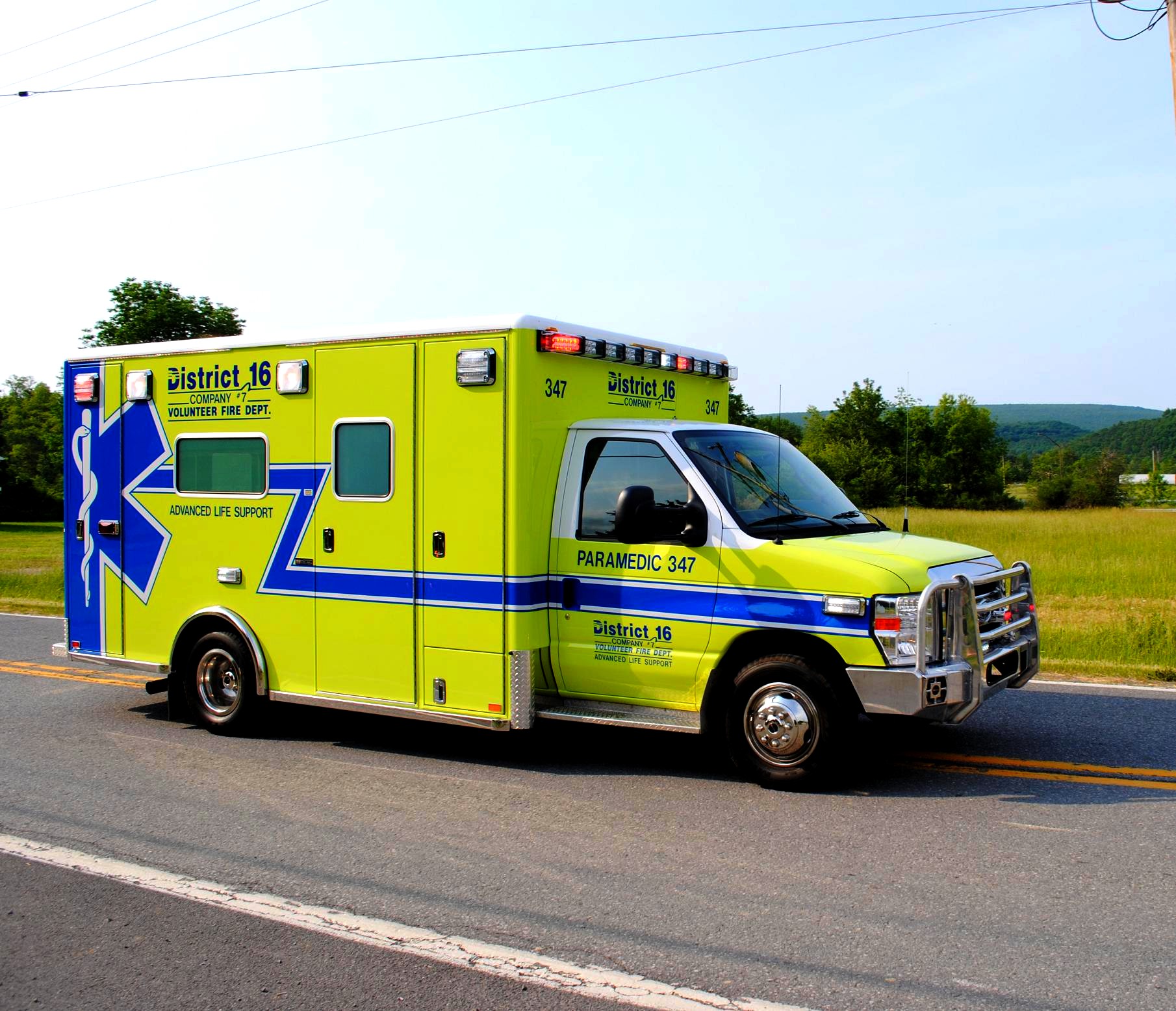 A lime-green paramedic ambulance driving on a two-lane road with green fields behind it.