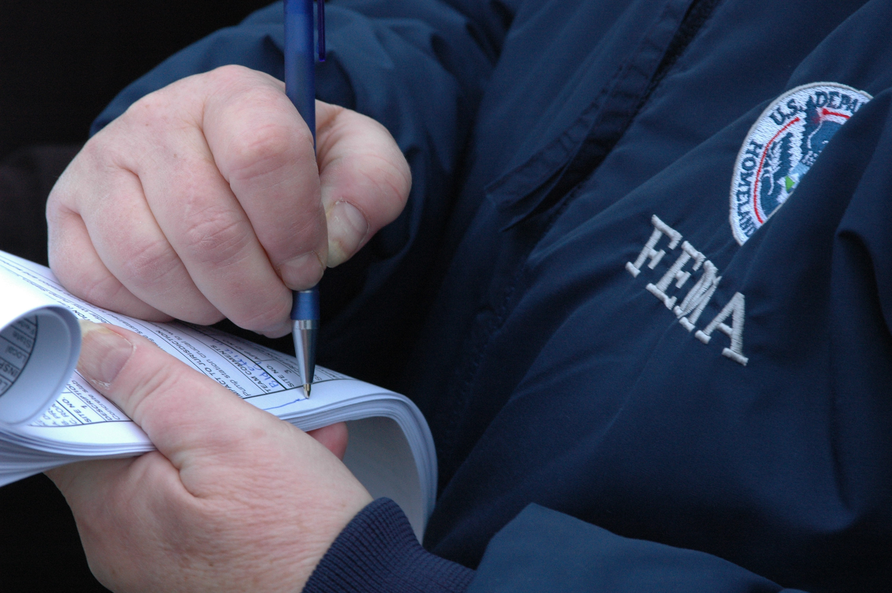 Person in FEMA jacket writing notes on a notepad with a blue pen.