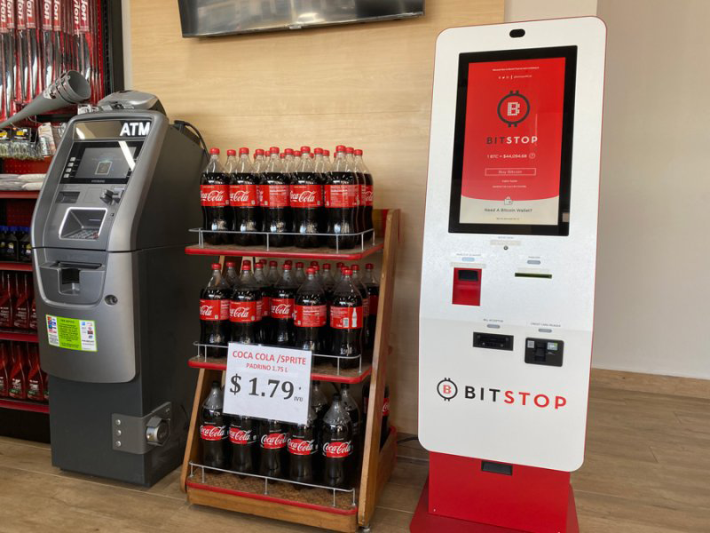 A red-and-white Bitcoin ATM kiosk stands next to a traditional ATM inside a store.