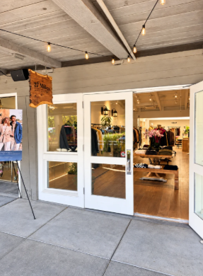 Open white-framed doors reveal clothing displays inside a 27 Miles Malibu boutique, beneath a covered walkway with string lights.