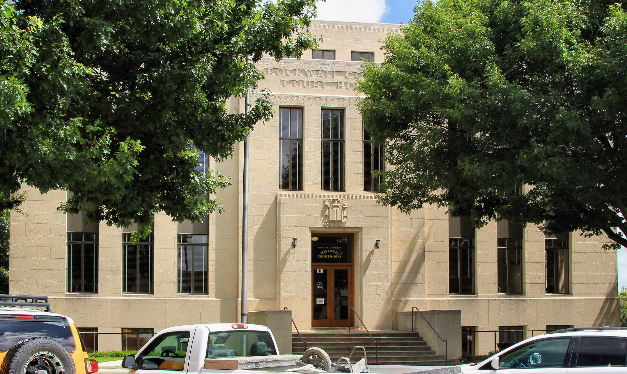 Tan stone courthouse building with ornate roofline details and large trees in front.