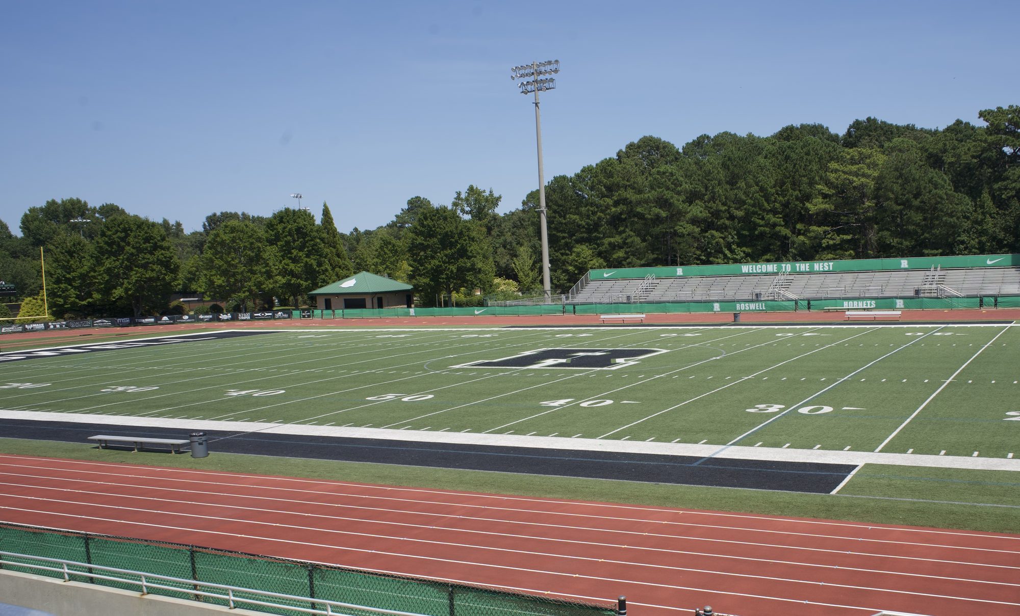 Empty high school football field with artificial turf, red running track, and a 'Welcome to the Nest' banner.