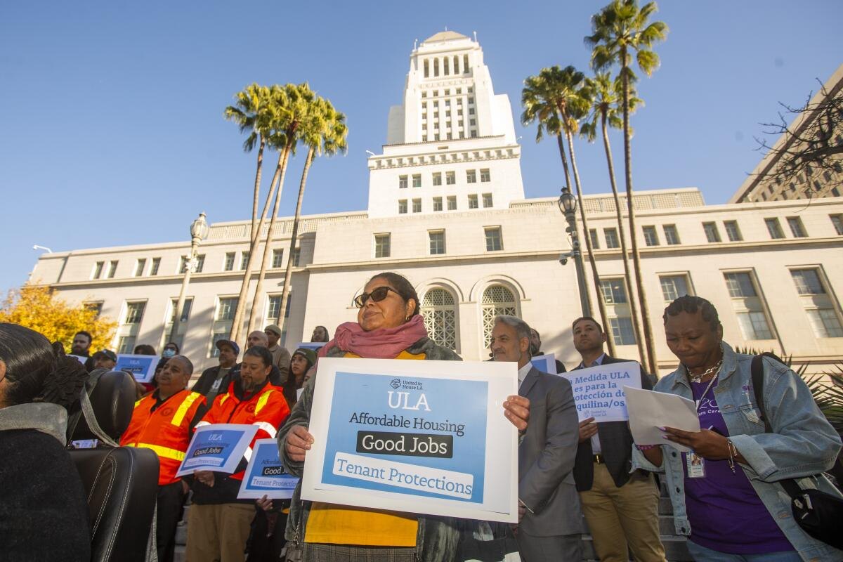 Woman holding a Measure ULA sign at a rally outside Los Angeles City Hall with palm trees in background.
