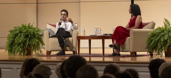 Two people seated in armchairs on a stage before an audience in an auditorium.