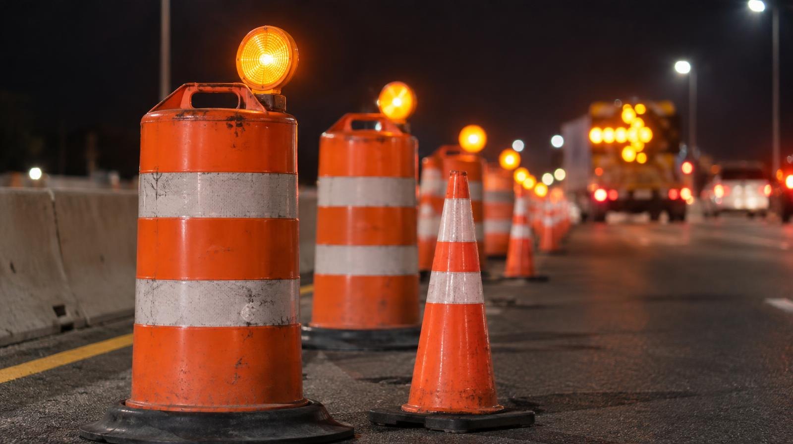 Orange traffic cones and amber-lit construction barrels along a closed highway lane at night.