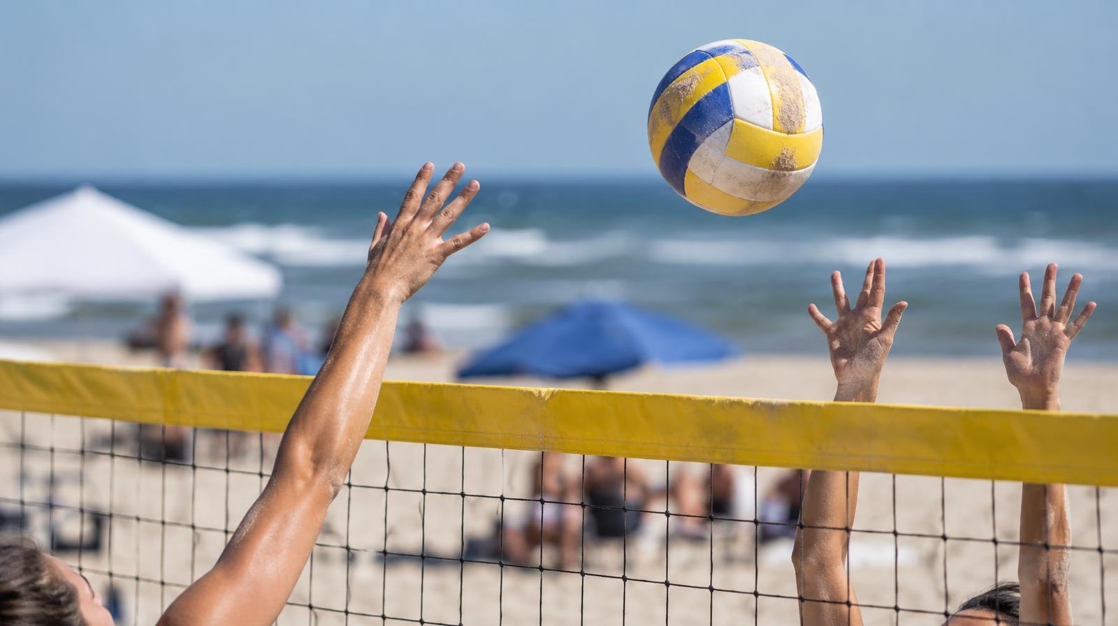 A beach volleyball in the air above a net with the ocean visible in the background.