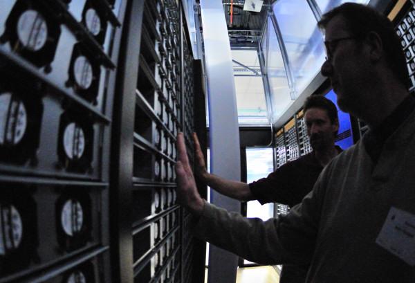 Two men standing between tall server racks in a blue-lit data center aisle.