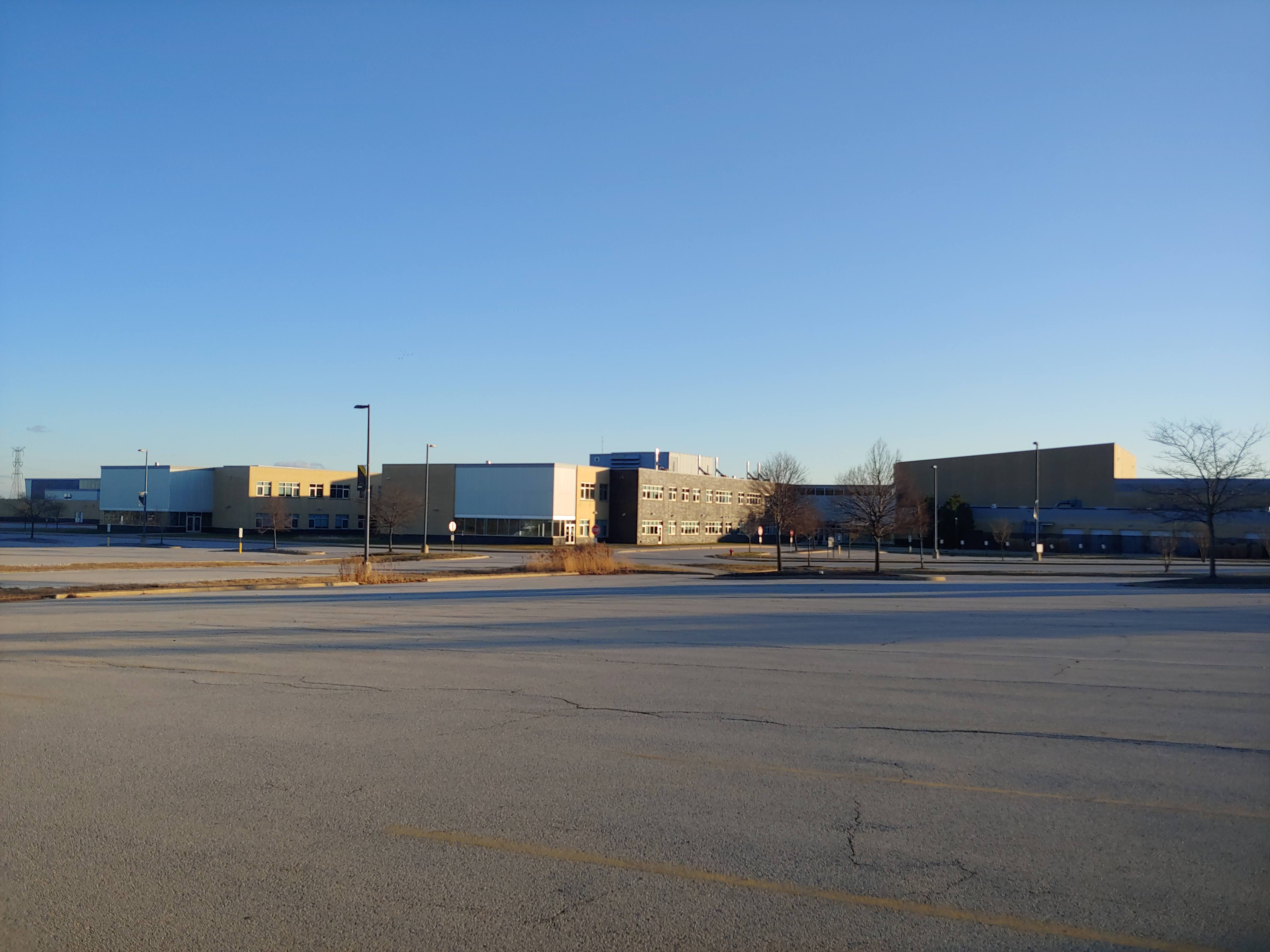 Wide exterior view of a large high school campus from an empty parking lot under a blue sky.