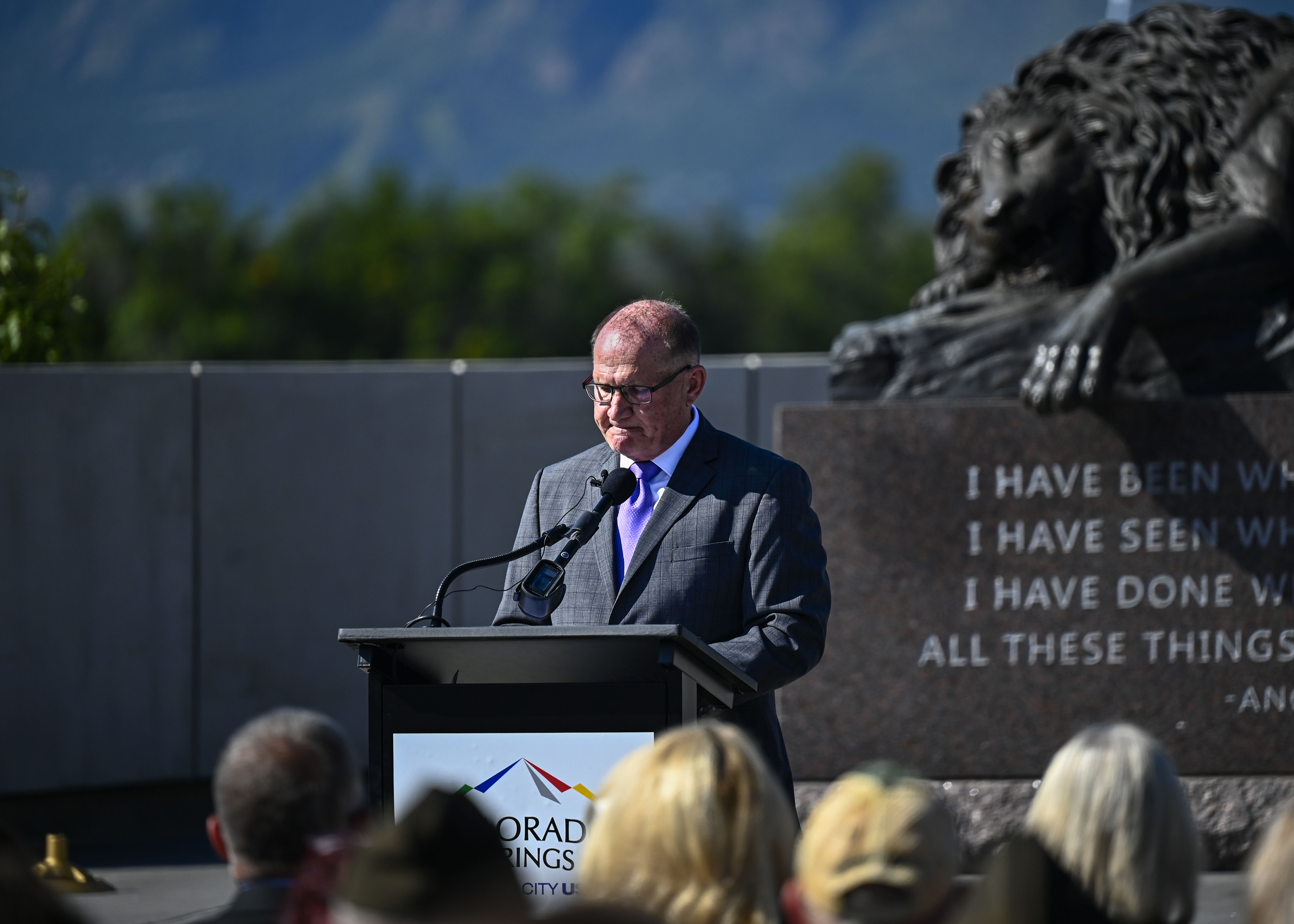 A man in a gray suit speaks at an outdoor podium at a 9/11 commemoration ceremony.