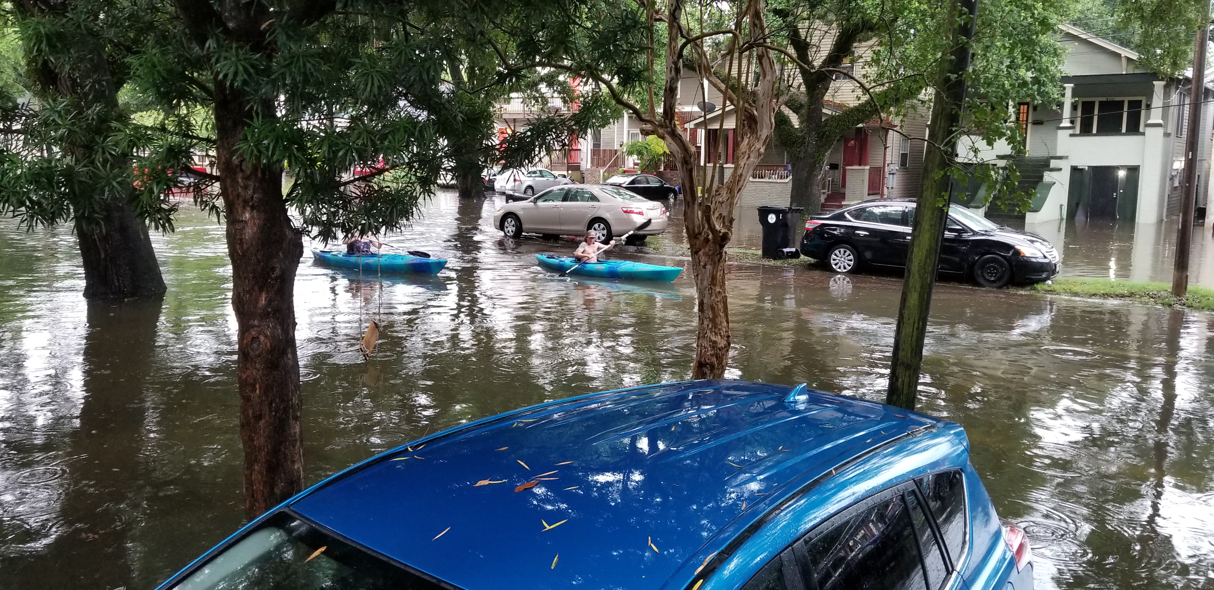 Two people kayak down a flooded residential street past partially submerged parked cars.