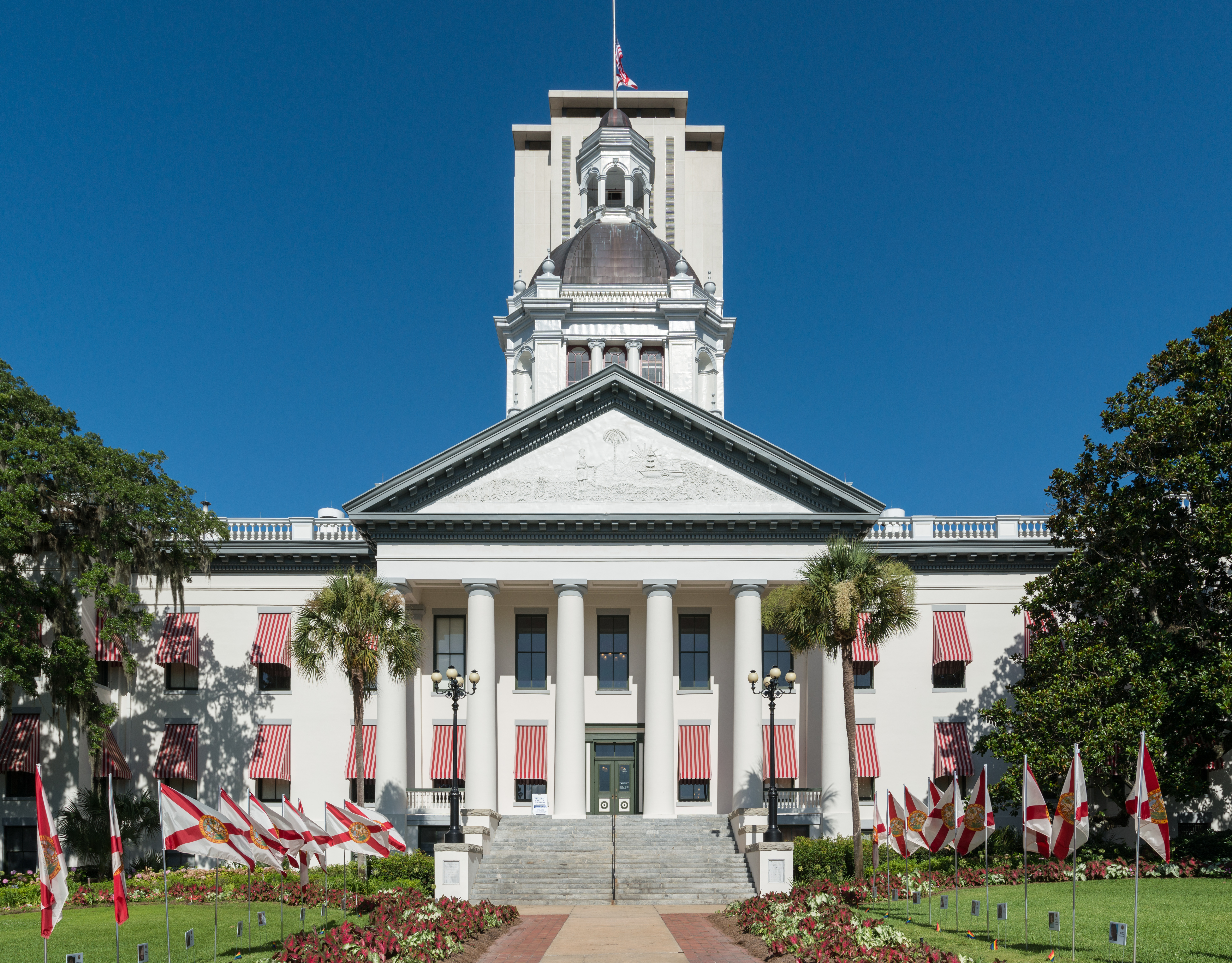 Historic domed Florida State Capitol building with palm trees and state flags, modern tower behind.