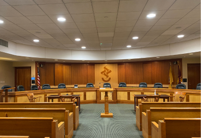 Empty municipal council chamber with raised dais, wooden paneling, and rows of public seating.
