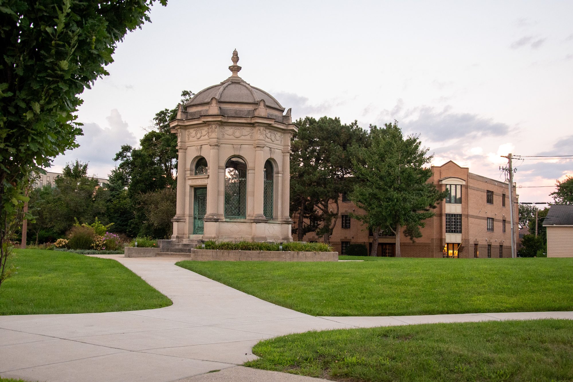 Stone neoclassical pavilion surrounded by mature trees in an Elmhurst park at dusk.