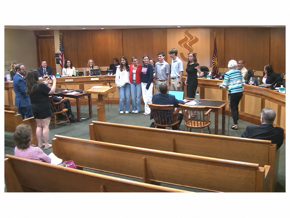 A group of high school students standing in a council chamber with council members seated at the dais behind them.
