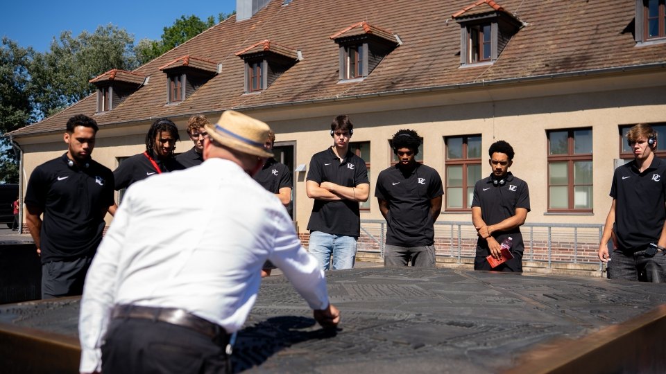 Group of young men in matching black shirts listening to a guide outdoors at a memorial site.