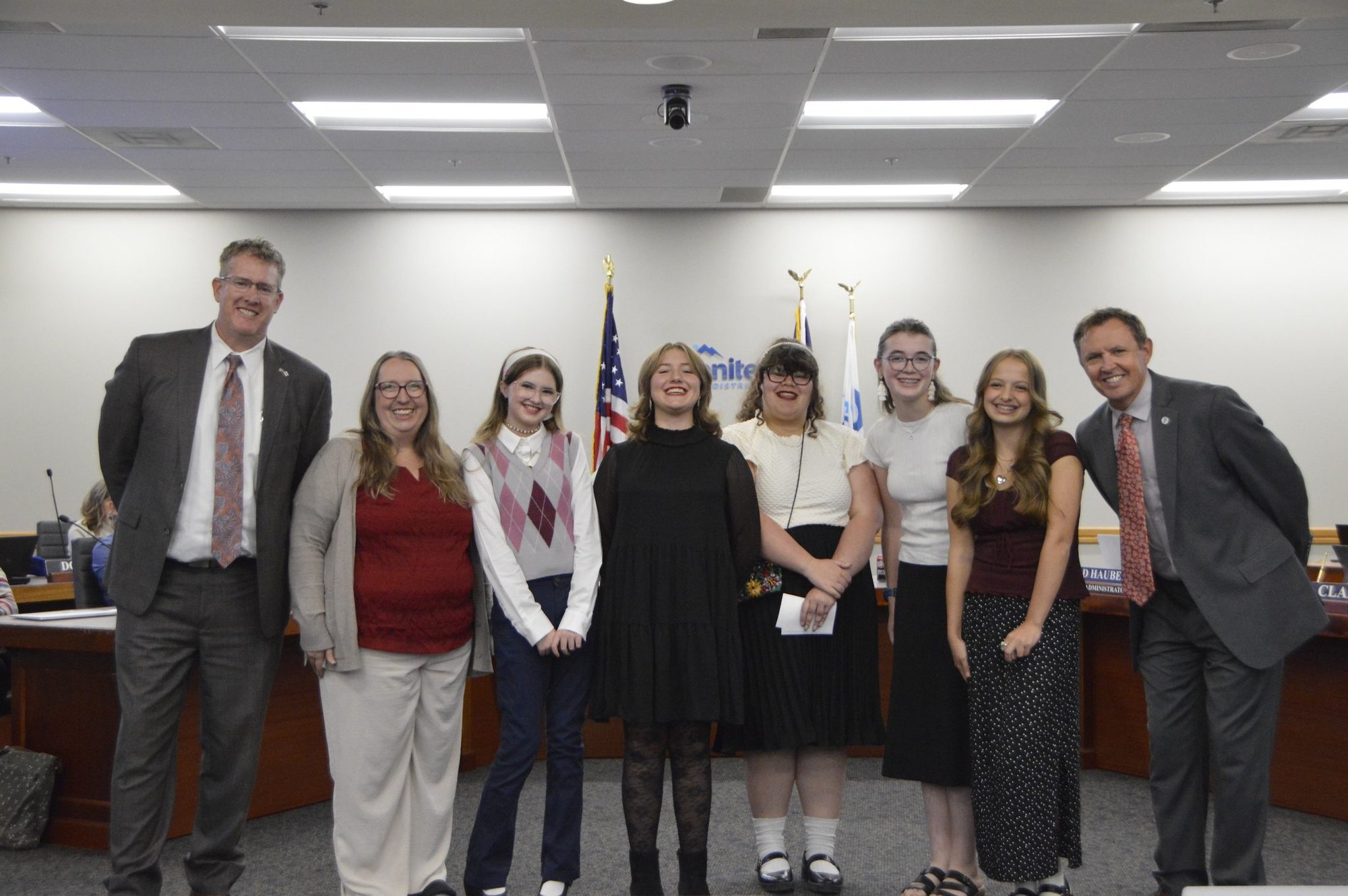 Group of students and adults posing in a formal school board meeting room with flags in the background.