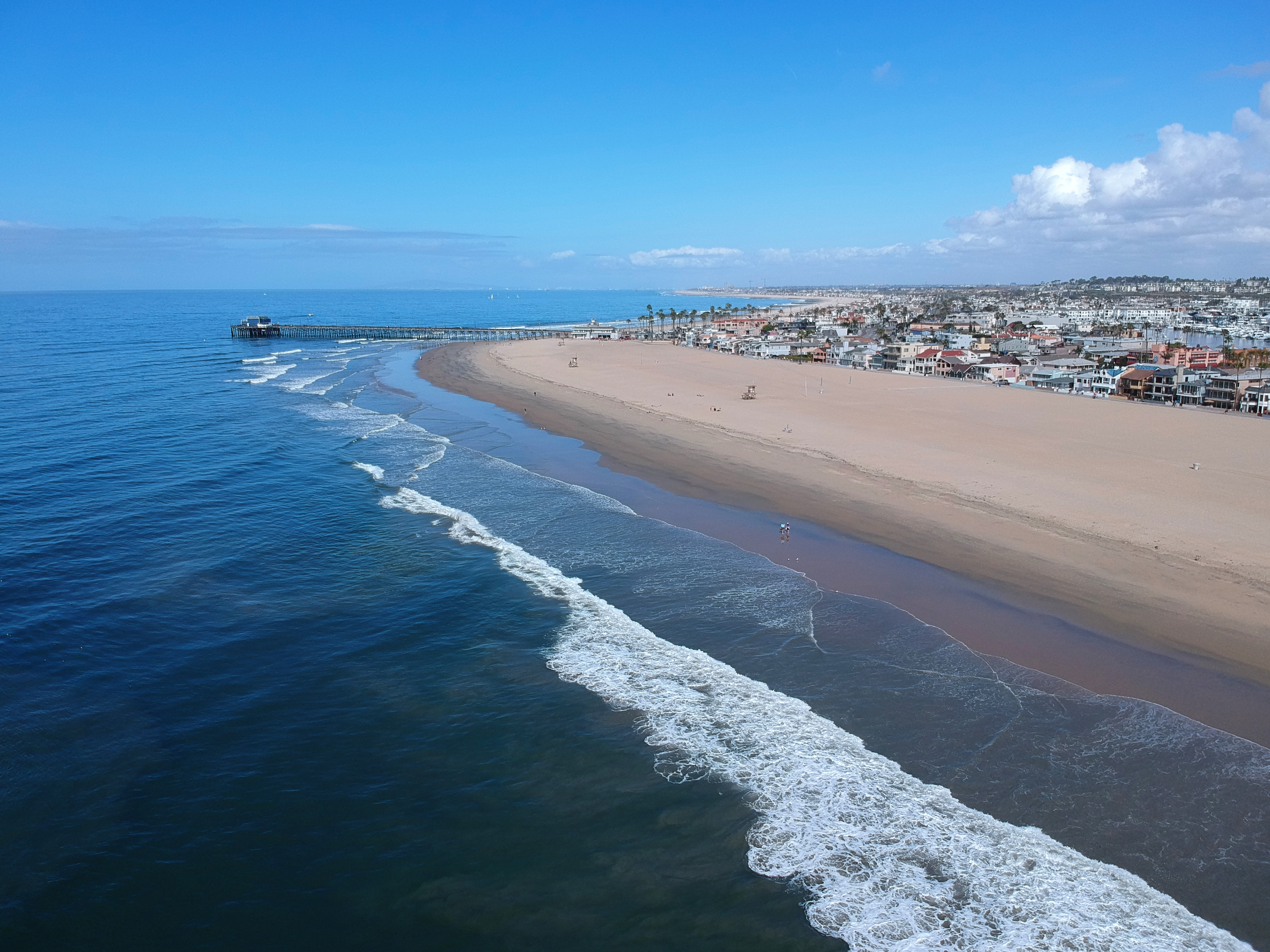 Aerial view of a sandy beach with breaking waves, a pier, and coastal homes.