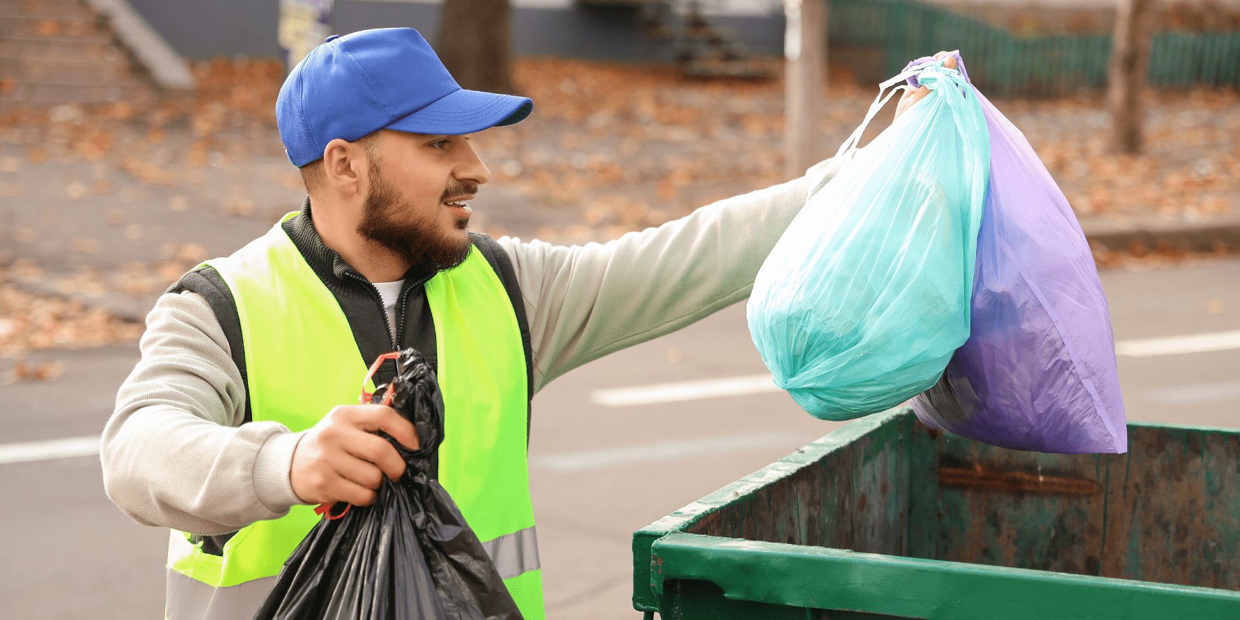 Worker in yellow safety vest drops trash bags into a green dumpster.