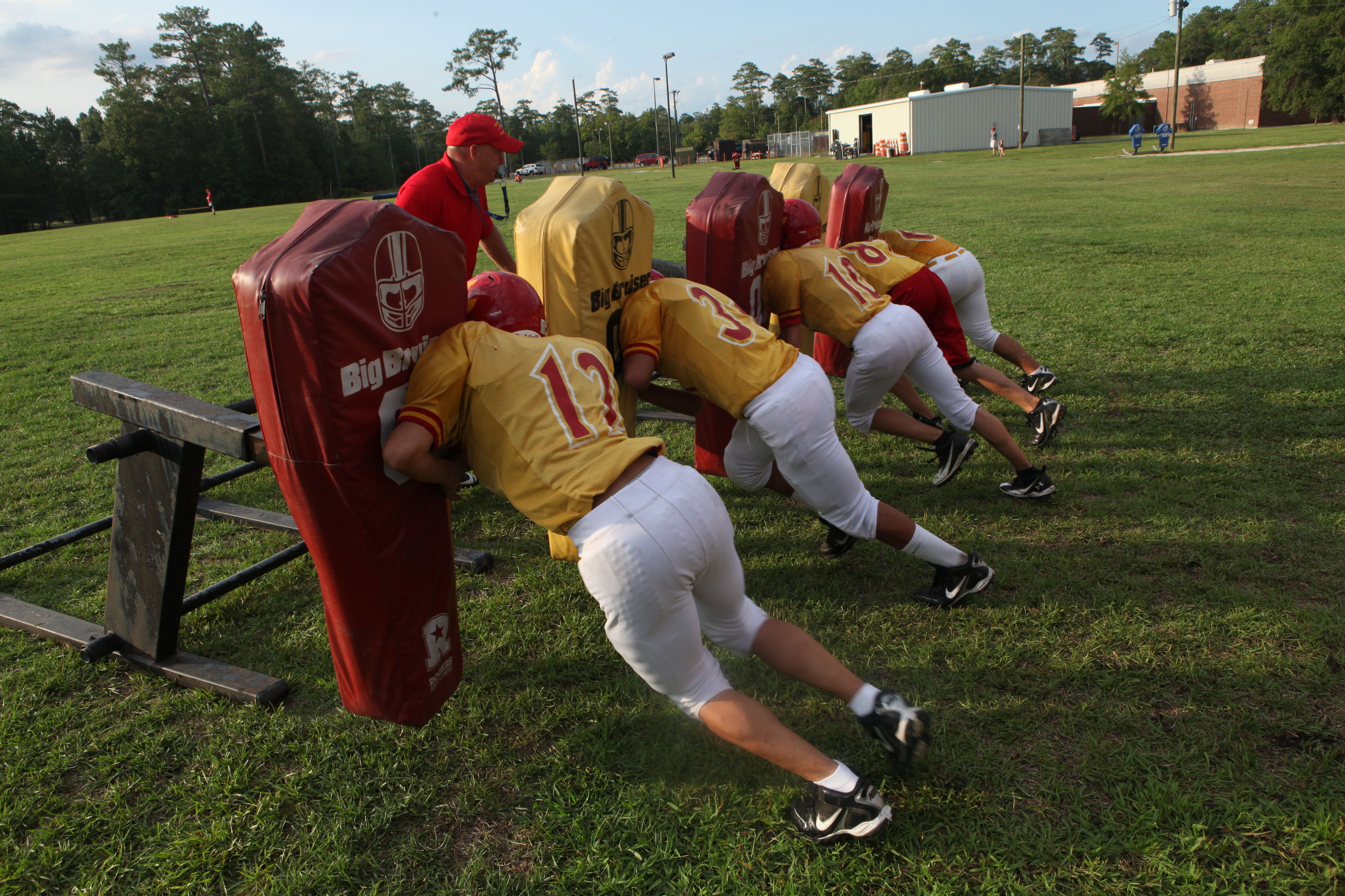 Football players in gold and white uniforms push a red blocking sled on a grass practice field.