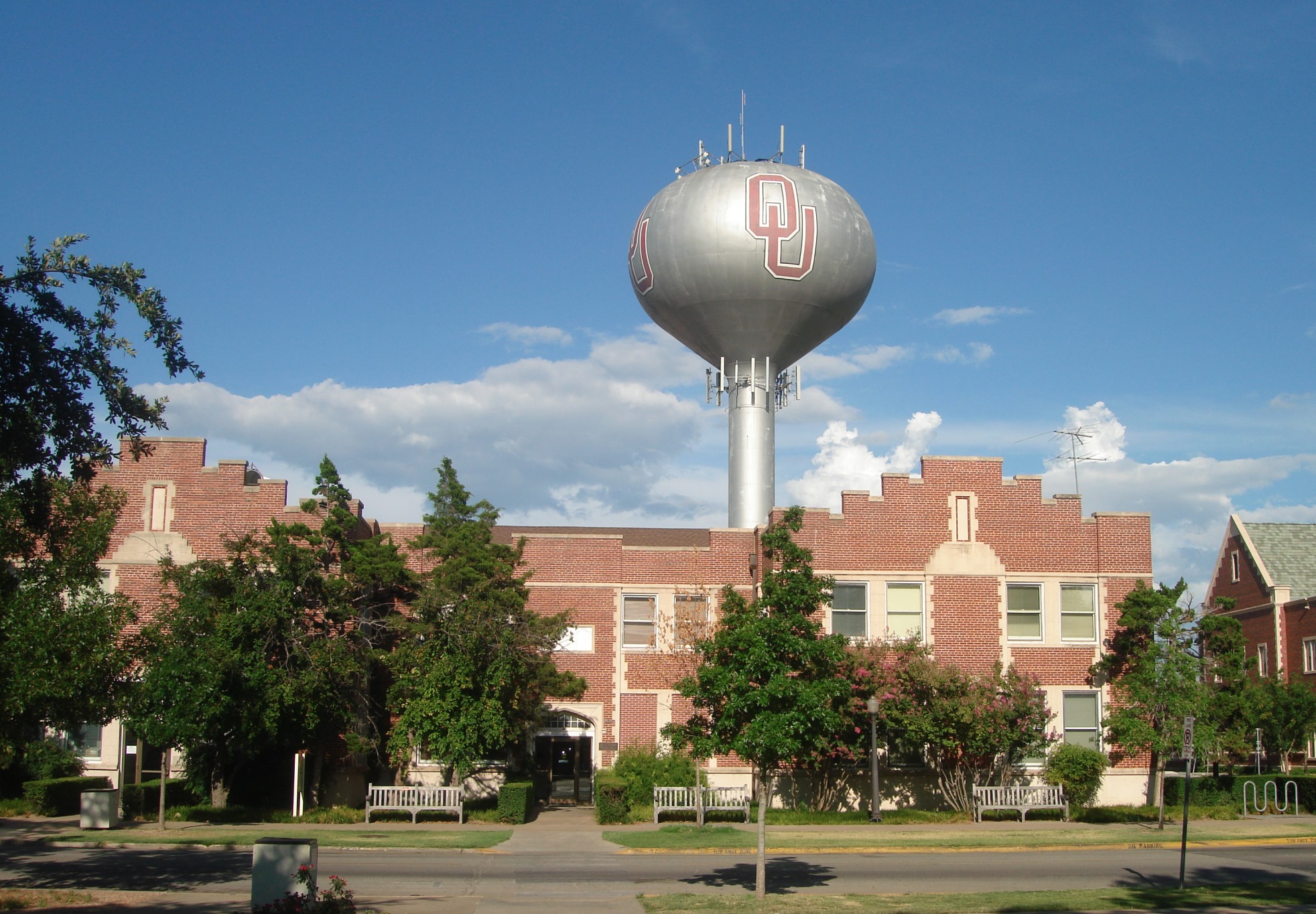 Silver spherical water tower with the OU interlocking logo above a brick building and trees.