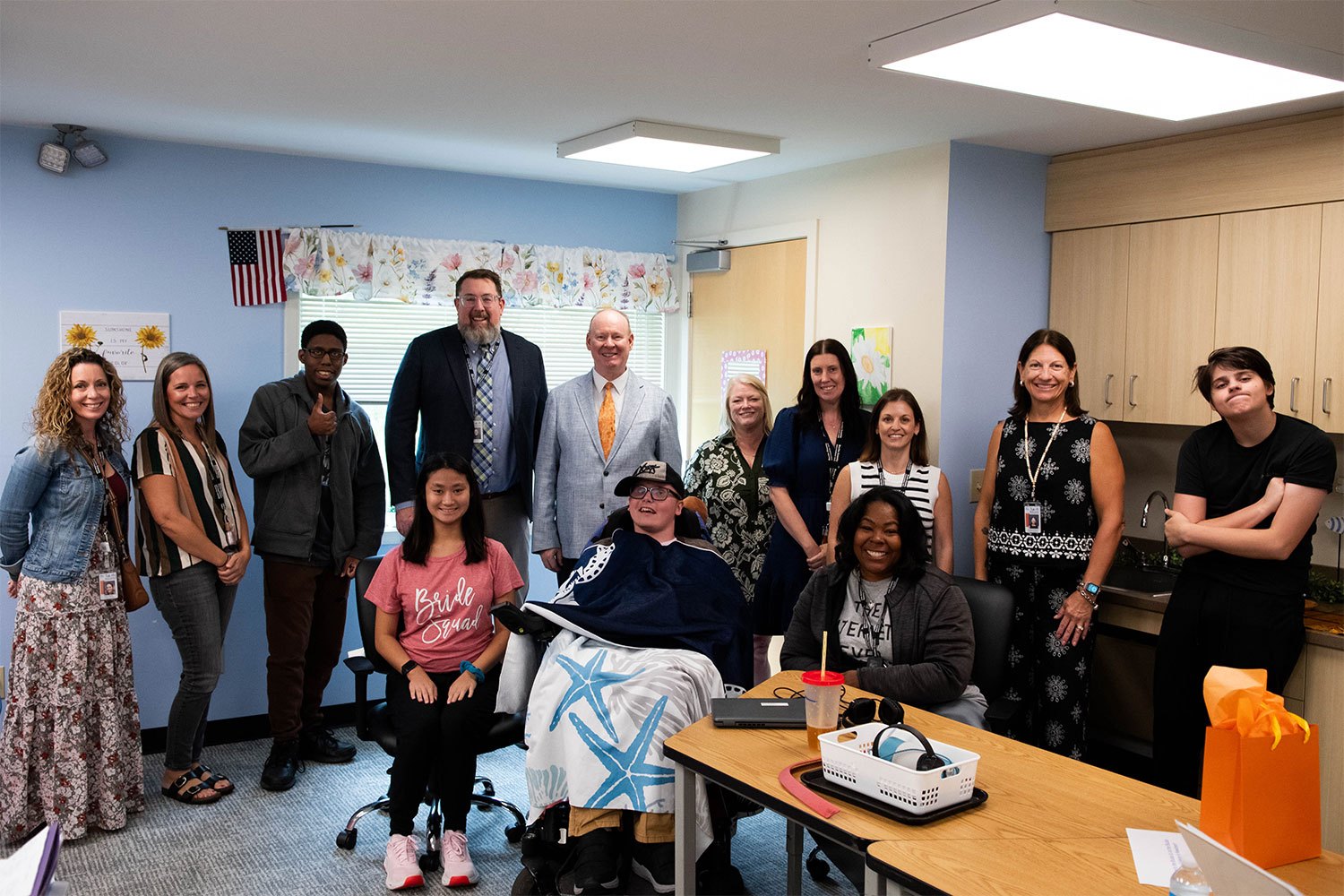 Group of adults posing indoors in an office room, one person seated in a wheelchair.