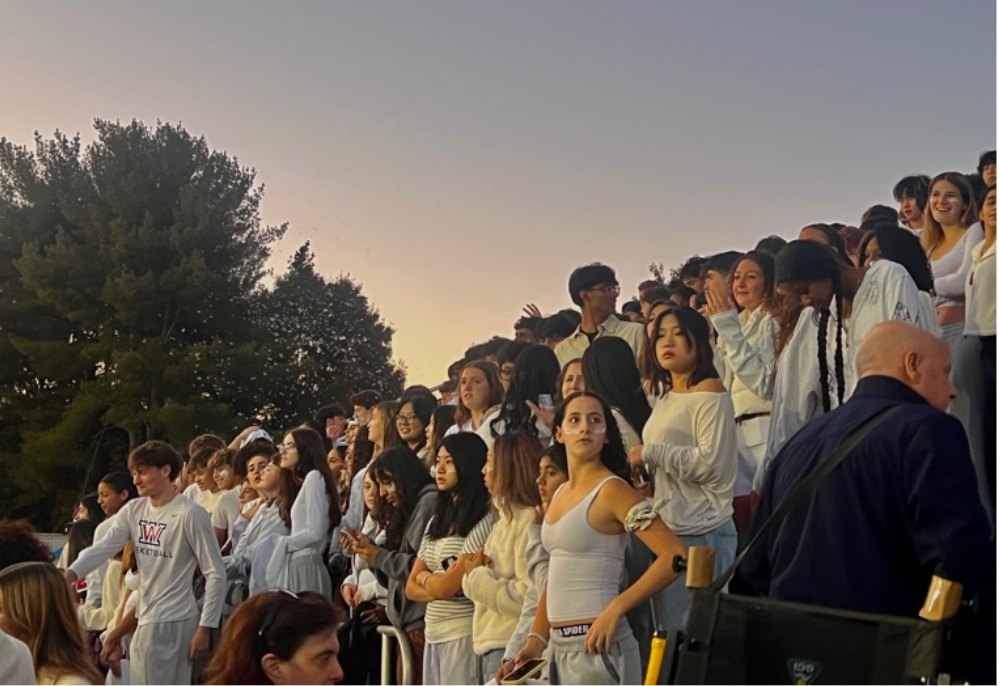 A large crowd of students in white clothing standing in bleachers at dusk.