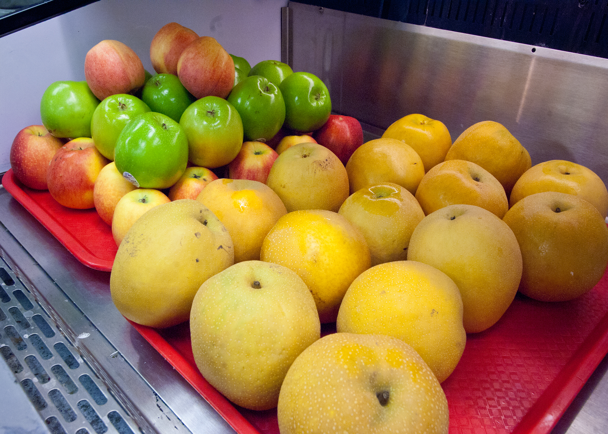 A red tray with fresh green apples, red apples, and Asian pears on a metal surface.