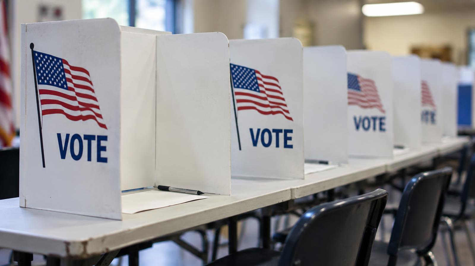 A row of white voting privacy booths on tables inside a brightly lit polling place.