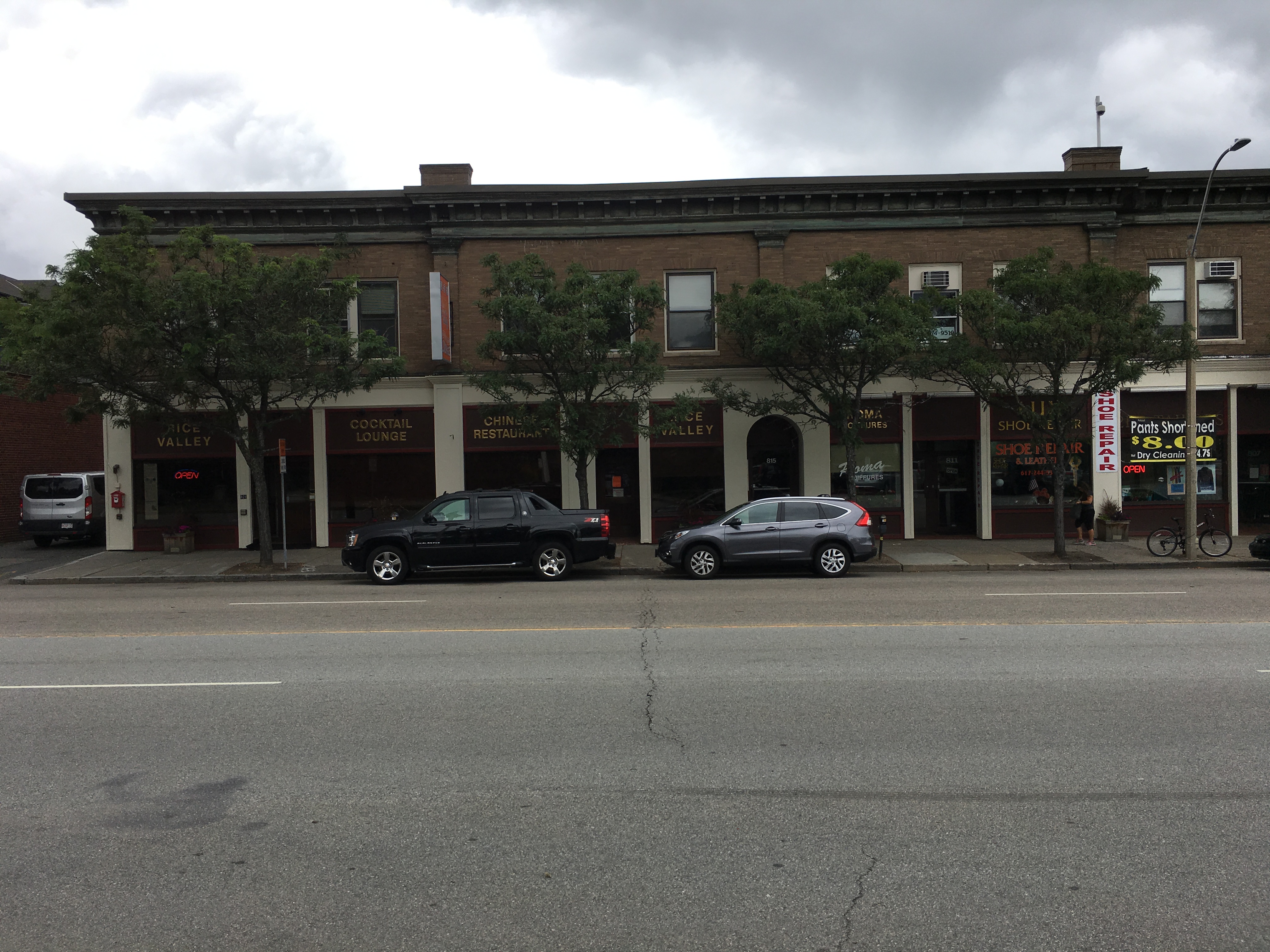 Brick storefronts and parked vehicles along a wide urban street in Newton, MA.