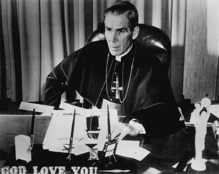 A bishop in vestments sits at a paper-covered desk with the words GOD LOVE YOU on its front.