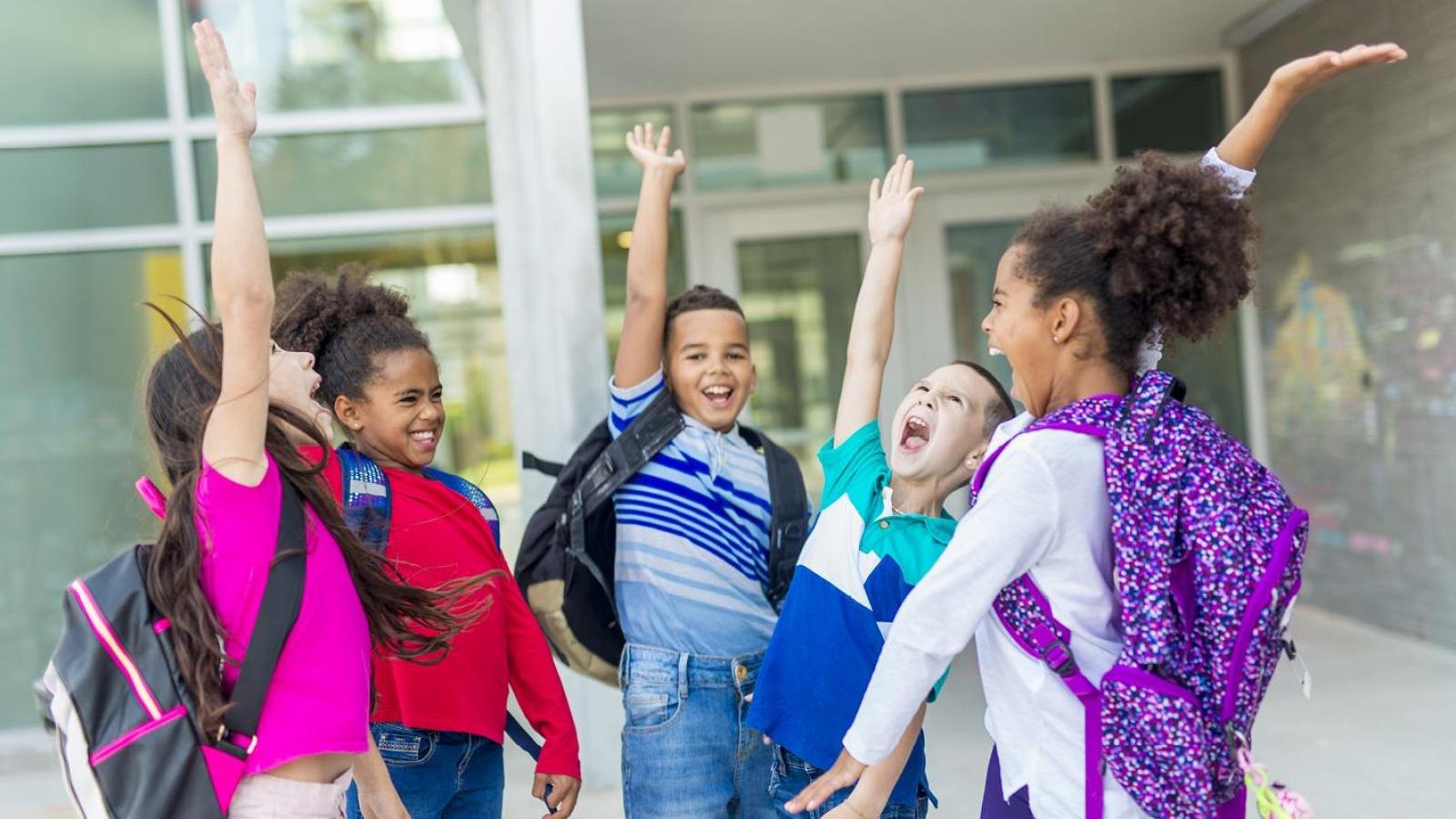 Five children wearing backpacks raise their arms in celebration outside a school entrance.