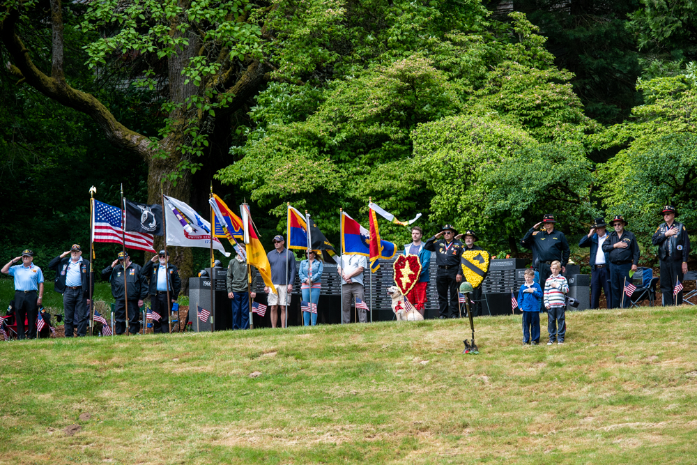 Veterans and civilians stand at attention with military flags at an outdoor memorial ceremony.