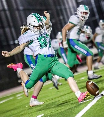Female football player in green Seahawks uniform kicking a football on a turf field at night.