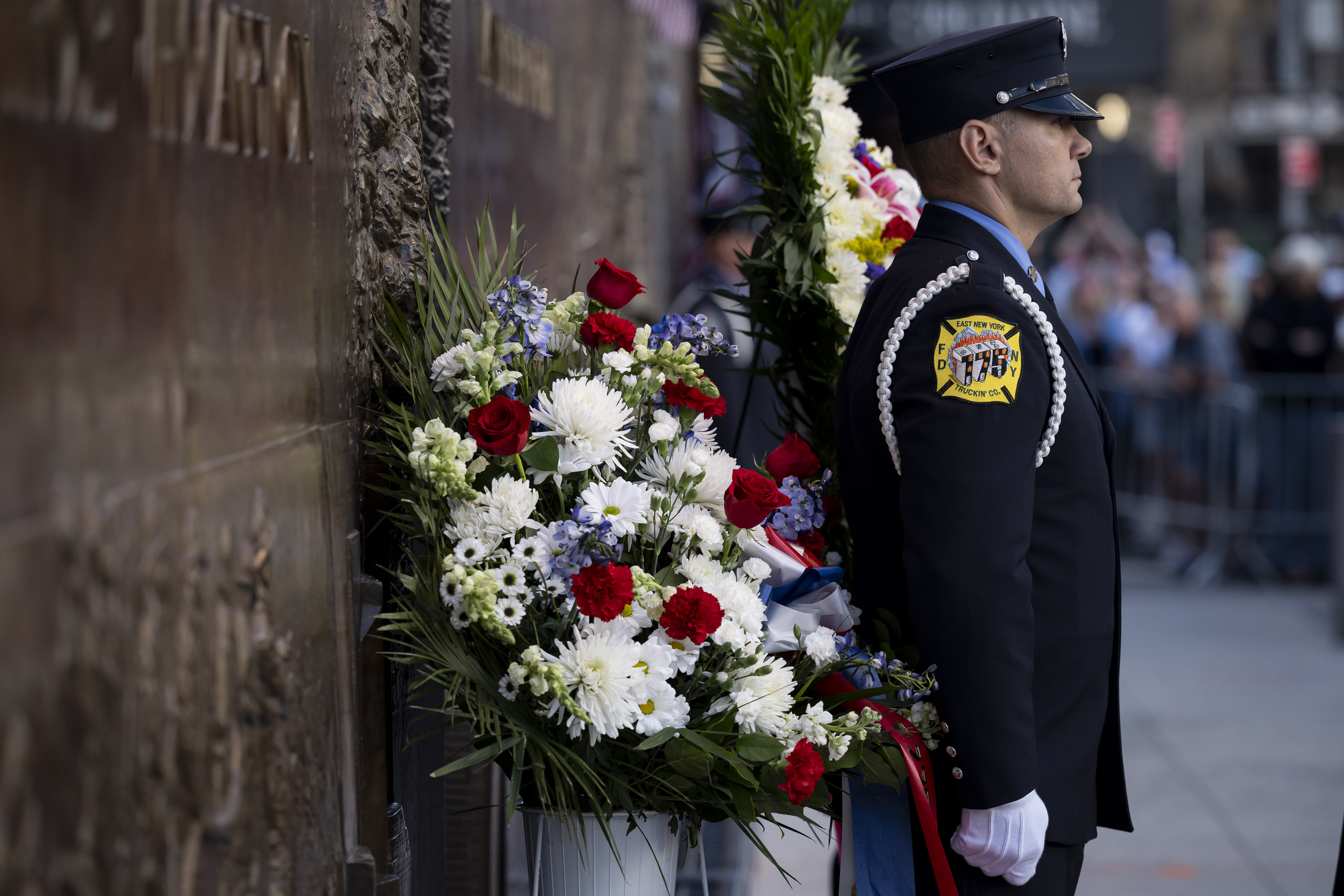Firefighter in white gloves and dress uniform stands beside a red, white, and blue floral wreath.